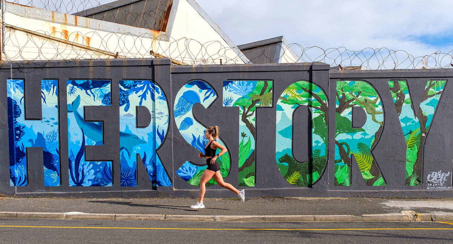 Coach Cords (Daniella Corder) in active wear running in front of a wall with graffiti "her story". 